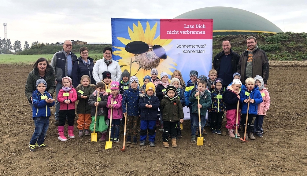 Foto: Die Kinder und Betreuenden des Kindergartens Lutzerath besuchen den Scholzehof Vergrößerung des Bildes für Eine Kindergartengruppe und 6 Erwachsene stehen vor einem Roll-up auf einem Ackerfeld. .