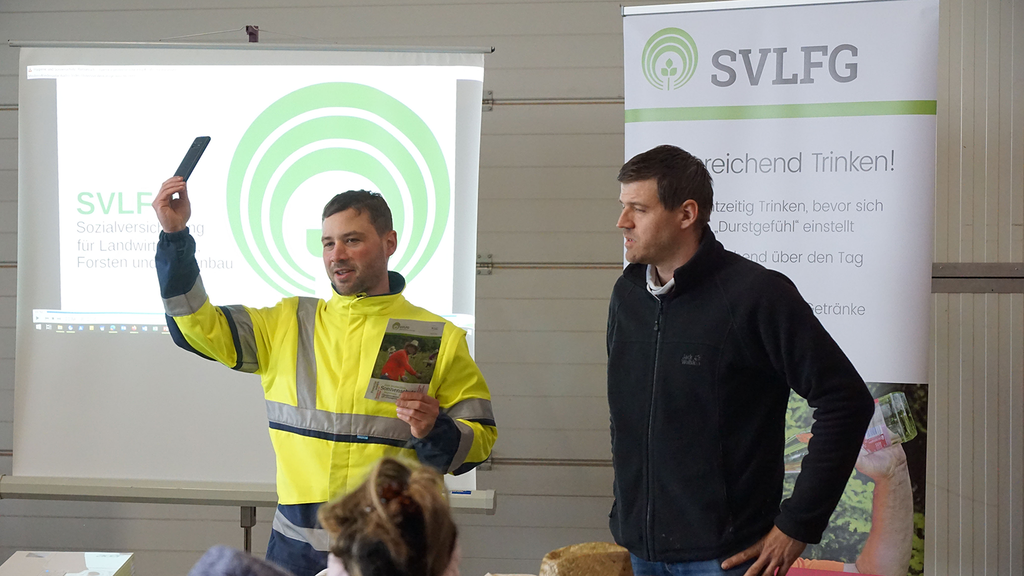 Web-App Erklärung Vergrößerung des Bildes für Zwei SVLFG-Präventionsexperten stehen vor einer Gruppe Saisonarbeitskräfte. Der linke Mann hält ein Smartphone in die Höhe und erklärt der Gruppe die WebApp..