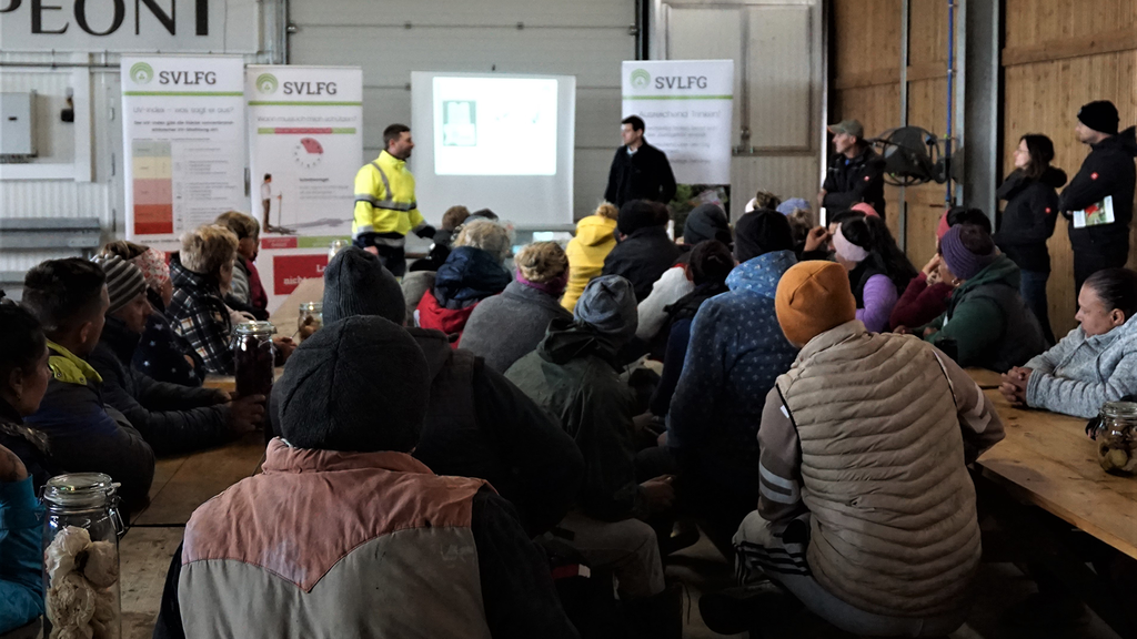 Gruppe der Teilnehmenden Vergrößerung des Bildes für Eine große Gruppe Saisonarbeitskräfte sitzt auf Holzbänken und hört einem Vortrag von Präventionsexperten zu. .