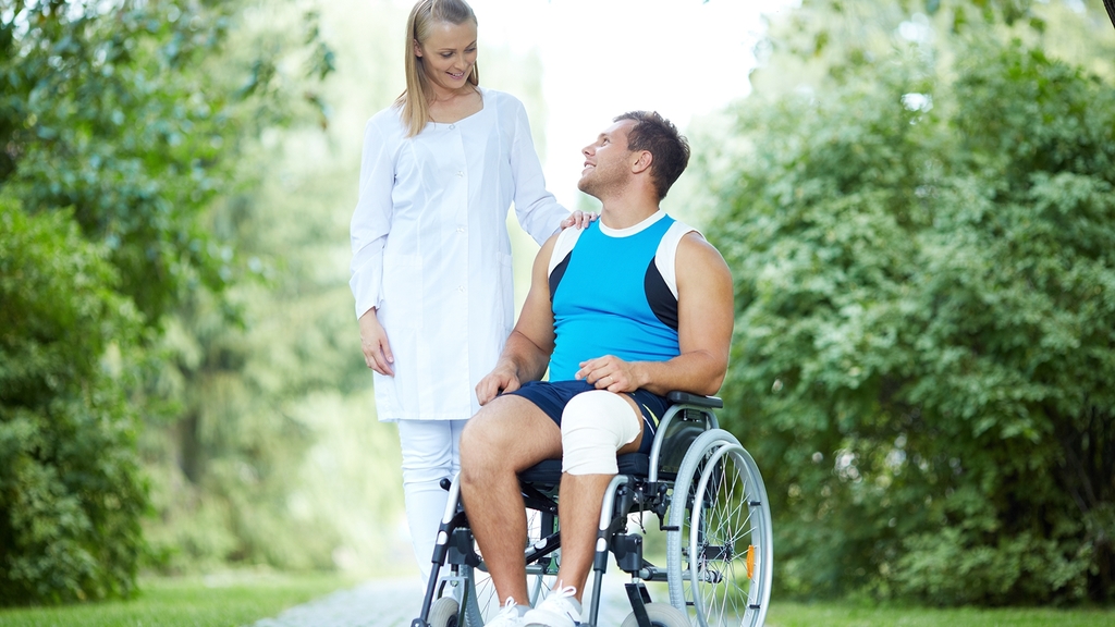 Vergrößerung des Bildes für Junger Mann mit Verband am Oberschenkel sitzt im Rollstuhl und schaut eine Krankenschwester an, die hinter ihm steht..